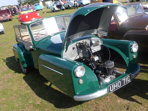 Green open three-wheeler with its bonnet tilted up showing the engine, on grass among microcars. Opens a larger view