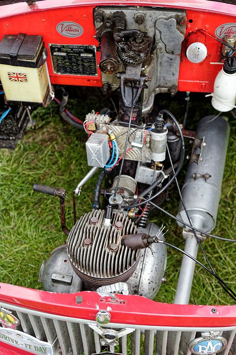 Looking down on a single-cylinder engine, exhaust and wiring above grass, the red grille at the bottom. Opens a larger view