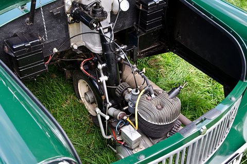 Finned single-cylinder engine and battery inside a green front compartment, grass beneath. Opens a larger view