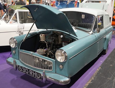 Pale blue three-wheeler with its bonnet raised over the engine, on a purple carpet beside a white van. Opens a larger view