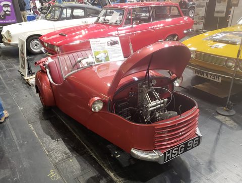Red three-wheeler with its whole nose tilted up, revealing the engine over the front wheel.