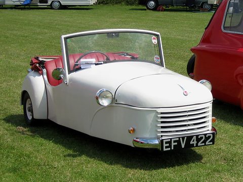White doorless three-wheeler with a slatted grille, red seats and a tall windscreen, parked on grass. Opens a larger view
