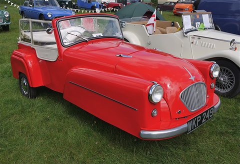 Red open three-wheeler with headlamps on its wings and white seats, on grass among cars. Opens a larger view