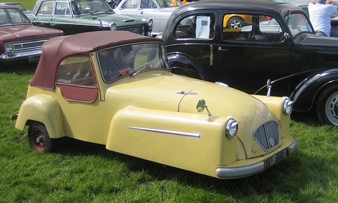 Yellow open three-wheeler with a maroon hood, headlamps on its front wings, parked on grass. Opens a larger view