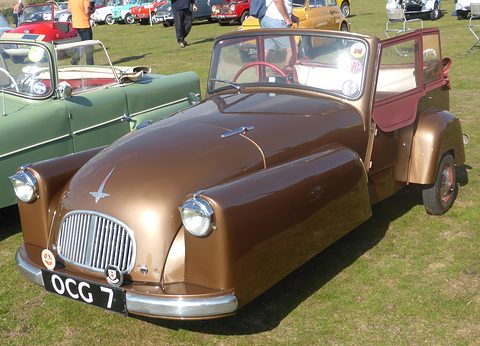 Metallic brown three-wheeler with an oval grille and badge bar, a green car beside it. Opens a larger view