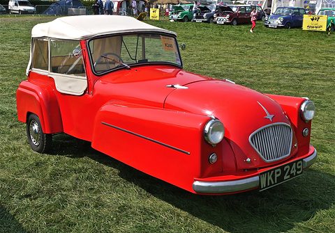 Red three-wheeler with a white soft top and chrome grille, parked on bright green grass. Opens a larger view