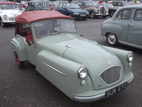 Pale green three-wheeler with a maroon hood and oval grille, parked on tarmac among classic cars. Opens a larger view