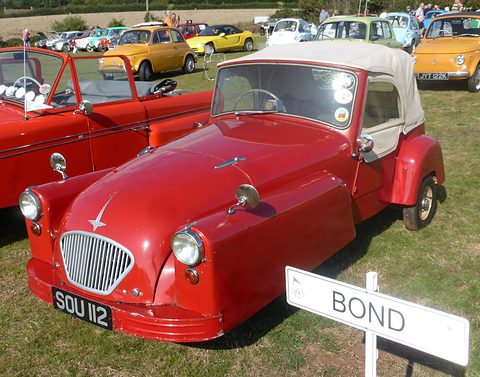 Red three-wheeler with a cream hood behind a white Bond signpost, on grass among other cars. Opens a larger view