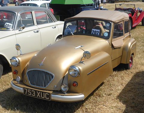 Gold three-wheeler with a beige hood and oval grille parked on dry grass. Opens a larger view