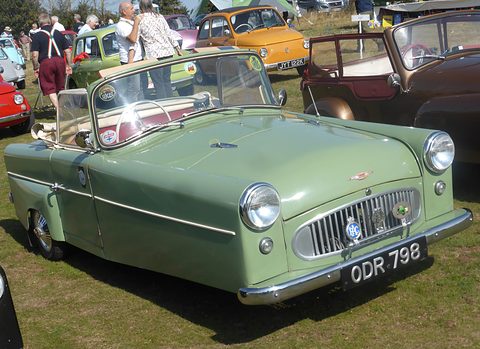 Pale green open three-wheeler with a full-width grille and chrome bumper, on grass among microcars. Opens a larger view