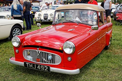 Red three-wheeler with a beige roof, chrome grille and bumper, visitors and cars behind it. Opens a larger view