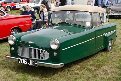 Green three-wheeler with a cream roof and chrome grille, parked on grass with visitors behind. Opens a larger view