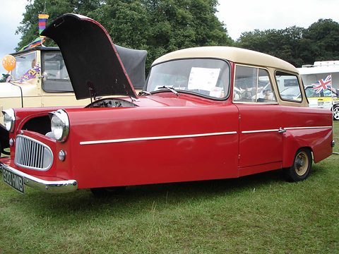 Red three-wheeler with a cream roof and raised bonnet, on grass under trees at a show. Opens a larger view