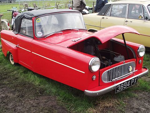 Red three-wheeler with a black hood and lifted bonnet, motorcycles and a cream car behind. Opens a larger view
