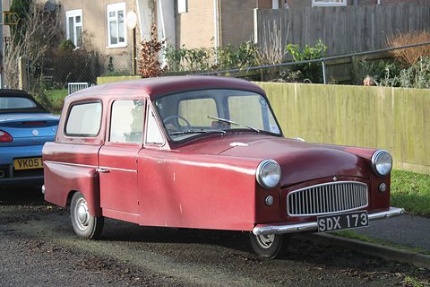 Two-tone red three-wheeler with a reverse-rake rear window, parked on gravel by a fence and houses. Opens a larger view