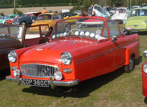Red open three-wheeler with a badge bar, RAC badge and twin lamps, on grass. Opens a larger view