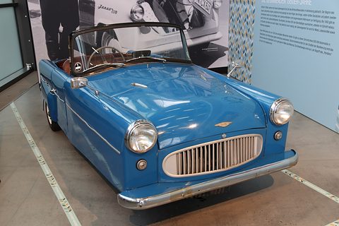 A small blue open three-wheeler with a wide slatted grille, chrome headlamp rims and a folded black hood frame. Opens a larger view