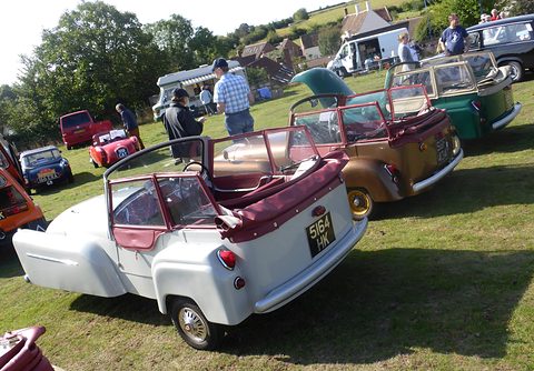 A white three-wheeler with a maroon hood seen from behind, a brown one beside it, on grass. Opens a larger view