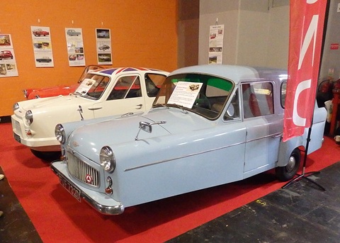 Pale blue three-wheeler saloon beside a cream one on a red carpet, posters on an orange wall. Opens a larger view