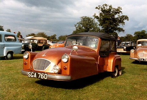 Copper-coloured three-wheeler with a black hood on grass, other small cars behind under a cloudy sky. Opens a larger view