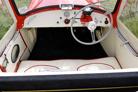 Looking down into a cream cockpit with a white three-spoke wheel, switches and a black floor mat. Opens a larger view