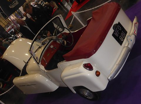 Cream three-wheeler seen from above and behind with red seats and a folded tonneau, on a show stand. Opens a larger view