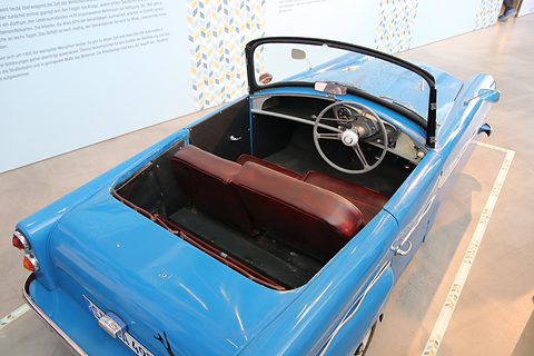 Looking into an open blue three-wheeler: a two-spoke steering wheel, a single dial and a red bench seat. Opens a larger view