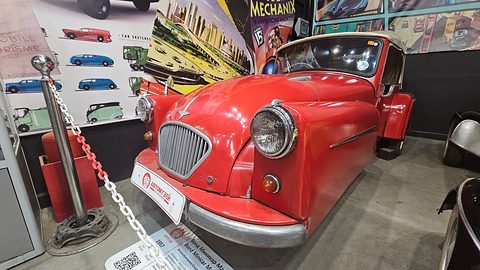 Red three-wheeler with an oval grille and wing-mounted headlamps, behind a chain barrier with posters on the wall. Opens a larger view