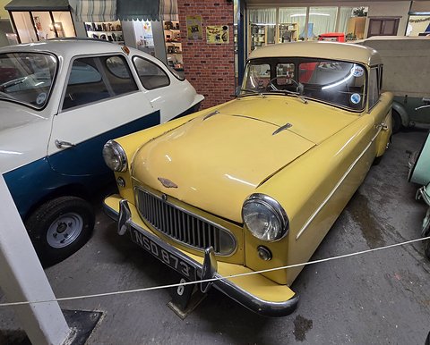 Yellow three-wheeler with a cream roof behind a rope, a brick wall and a white car beside it. Opens a larger view