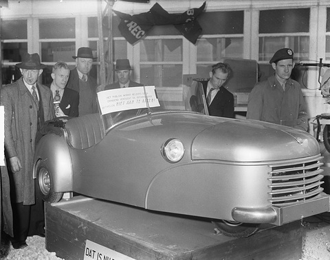 Black-and-white: a doorless silver three-wheeler on a plinth, men in hats and coats looking on.