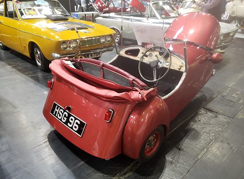 Red open three-wheeler seen from behind with its hood folded, other classic cars around it. Opens a larger view