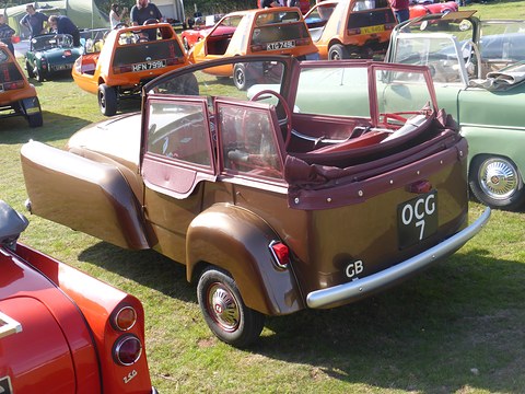 Brown three-wheeler with open doors, a hood frame and tonneau, parked on grass among microcars. Opens a larger view