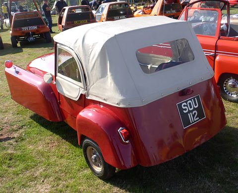Red three-wheeler with a cream hood and rear window seen from behind, door open, on grass. Opens a larger view