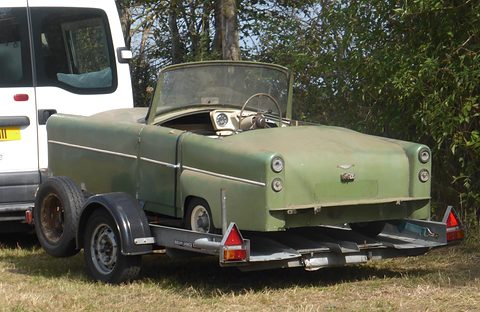 Weathered pale green three-wheeler with flared rear wings sitting on a trailer by a hedge. Opens a larger view