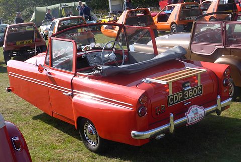 Red open three-wheeler with a wooden luggage rack on its tail, on grass among other cars. Opens a larger view