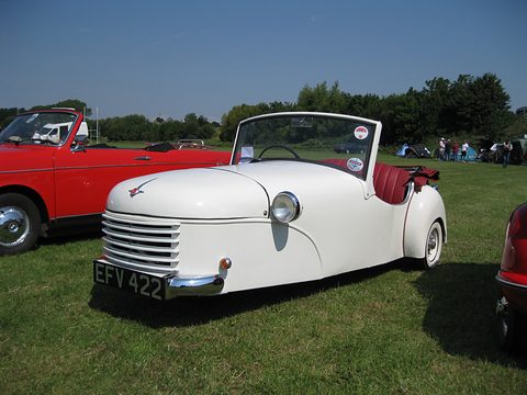 White three-wheeler seen from the side on a show field, a red sports car behind it.