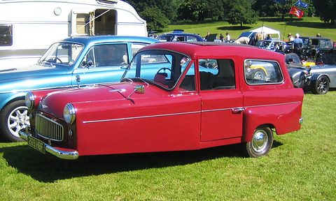 Red three-wheeler with a long roof and side windows in profile, parked among cars on grass. Opens a larger view