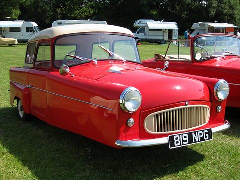 Red three-wheeler with a cream roof and side windows, parked on grass beside a red open car. Opens a larger view