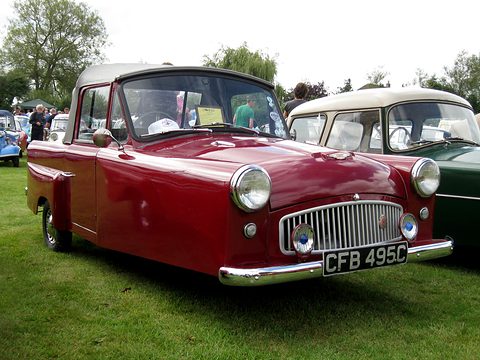 Dark red three-wheeler with a black top and chrome grille, on grass beside a cream car. Opens a larger view