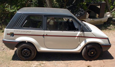 Side view of a beige and grey small two-box car with a BR-800 side script, louvred rear pillar and red stripe, parked on dirt by trees. Opens a larger view