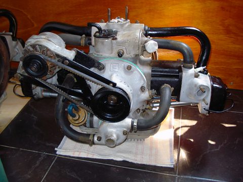 A small flat-twin engine with black pipes and a belt-driven alternator, standing on a dark polished table.