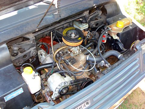 Engine bay under a raised grey bonnet: a flat-twin with an orange and black air cleaner, a coolant bottle and wiring. Opens a larger view