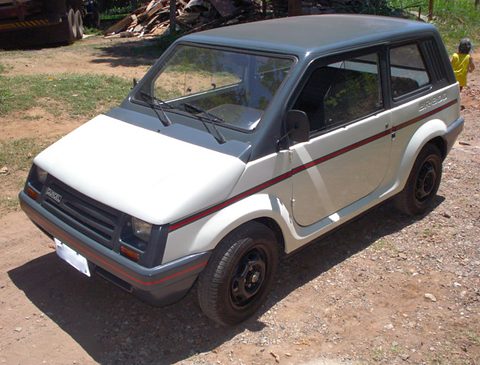 Front quarter of a beige small two-box car with a grey roof, grey bumpers and a red stripe, parked on bare earth. Opens a larger view