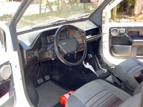 Small car cabin through the open door: black steering wheel, a plain dashboard with square dials, grey seats and a red fire extinguisher. Opens a larger view