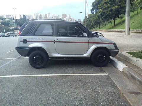 Side view of a silver and grey small two-box car with flat glass and a red side stripe, parked across a kerb, tower blocks behind. Opens a larger view