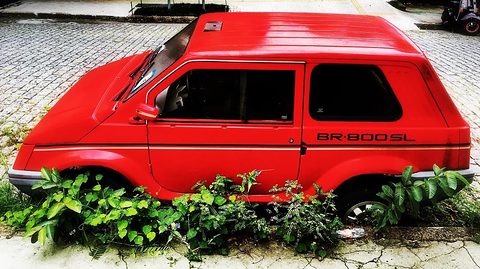 Red small two-box car with a BR-800 SL script on its flank, parked over a strip of plants on a cobbled street. Opens a larger view