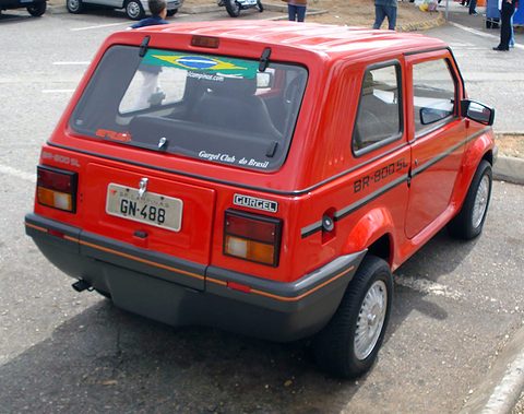 Rear quarter of a red small two-box car with a Brazilian flag sticker on its rear window and a grey bumper, on a car park. Opens a larger view