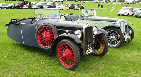 A grey and black three-wheeler with red wire wheels parked on grass beside a pale green sports car. Opens a larger view