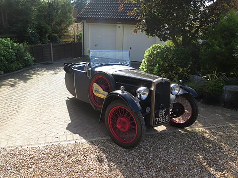A black open three-wheeler with red wire wheels and a chrome radiator on a gravel drive before a garage. Opens a larger view