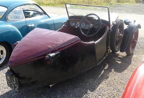 Rear three-quarter view of a maroon and black three-wheeler with a pointed covered tail and a spare wheel. Opens a larger view